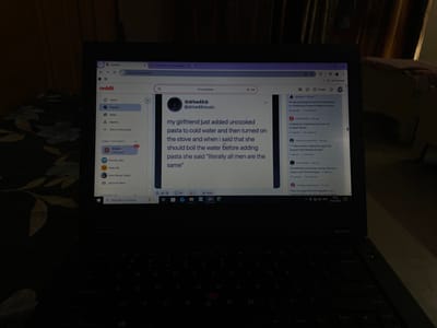 The scene depicts a dimly lit indoor environment in Noida, India, centered on an open laptop. The laptop, dark in color, rests on an unseen surface. To the immediate left, a dark fabric with a light floral or leaf pattern is visible. The background is out of focus, showing blurred forms of furniture, possibly a bed with a light-colored headboard or bedding.
The laptop screen is illuminated, displaying a Reddit webpage. The left panel of the Reddit interface shows navigation options ("Home," "Popular," "News," "Explore," "Start a community") and several subreddits under "GAMES ON REDDIT" (e.g., "r/BikiniBottomTwitter," "r/plunder_isles"). The central content area prominently features a post by user "@drive45music," which reads: "my girlfriend just added uncooked pasta to cold water and then turned on the stove and when i said that she should boil the water before adding pasta she said 'literally all men are the same'". The right panel displays a list of other Reddit posts, with partially visible titles such as "...GF was planning to meet his new friend..." and "Looking for Angel Investors for my Sneaker Brand." The Windows taskbar at the bottom of the screen displays the "Type here to search" bar, several application icons, and the system clock showing "19:33 16-10-202X". No individuals are visible in the image.