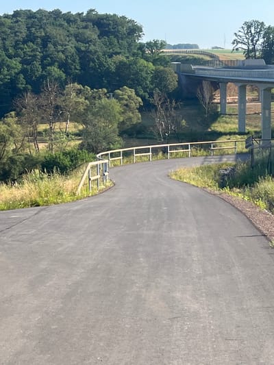 The media file shows a paved path curving to the right, with a grassy embankment on the left. The embankment is bordered by a metal railing. Beyond the embankment, there is a dense forest on the left and a bridge structure on the right. The bridge is elevated on concrete pillars and appears to be a highway or overpass. In the background, there is a field with what looks like a wind turbine and some scattered trees. The sky is clear and blue, suggesting a sunny day. The scene is outdoors and appears to be a park or recreational area, possibly near a highway. There are no people visible in the image. The time of day appears to be midday, given the bright lighting and clear sky. The overall impression is one of a natural landscape with some man-made infrastructure.