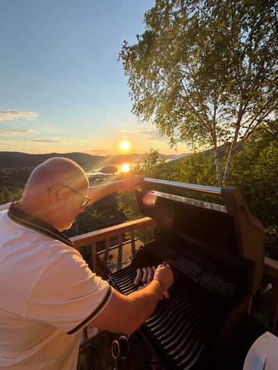 A man with glasses and a light-colored polo shirt is grilling sausages on a barbecue on a wooden deck during sunset. The sun is setting over a body of water, casting a warm glow on the landscape of hills and trees. A birch tree with green leaves is visible to the right of the frame. The setting appears to be a residential area in the city of Mont-Tremblant, Canada, given the natural mountainous terrain and the presence of a lake or large river. The man is leaning over the grill, his right hand reaching to adjust a sausage. The grill is open, and several sausages are visible on the grates. The sky is clear with a few scattered clouds.