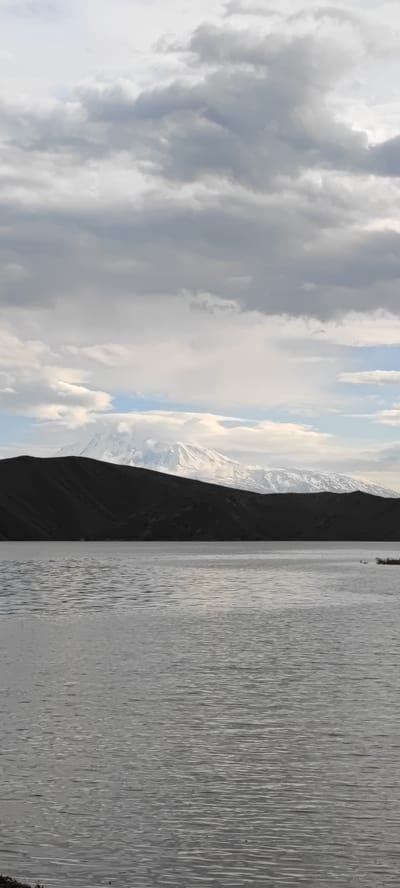 A vertical shot shows a still body of water under a cloudy sky with a dark, low-lying landmass in the middle ground and a snow-capped mountain range in the background.

The water's surface is rippled, reflecting light and creating a sense of calm. In the middle ground, a dark, seemingly barren landmass stretches horizontally across the frame, its shape uneven and broken. Behind this landmass, the snow-capped mountains rise, their peaks visible despite the cloud cover.

The sky is filled with grey clouds of varying density. This suggests overcast conditions.

The location is Lanjazat, Armenia, due to the presence of Lake Sev and the Gegham Mountains.