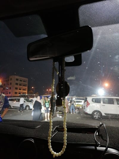 The image is a nighttime, interior shot taken from a car in Inezgane, Morocco. The windshield is visible, with water droplets and a reflective texture from a sun visor. A rearview mirror hangs from the top center of the windshield, along with a string of prayer beads. Outside the car, a group of people are visible in what appears to be a parking lot or street. Some are walking, and one person is wearing a green t-shirt with the number "33". In the background, there is a multi-story building and several white vehicles. Streetlights cast an orange glow on the scene. The overall lighting is dim, with artificial lights from the street and buildings illuminating the outdoor environment.