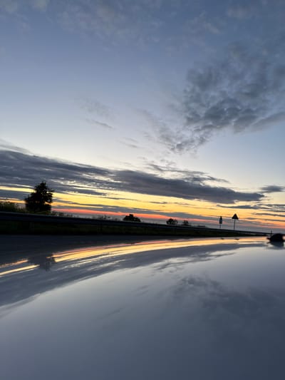 This image captures a sunset over a road in Paldiski, Estonia. The sky transitions from a pale blue at the top to a vibrant band of orange and red near the horizon, with streaks of grey clouds scattered throughout. In the distance, a line of trees marks the edge of the landscape. A highway guardrail runs along the side of the road. The surface of the road, likely wet, reflects the colors of the sunset and the sky, creating a mirror-like effect. Two traffic signs are visible on the right side of the road. The overall scene is serene and atmospheric, showcasing the beauty of a coastal sunset.