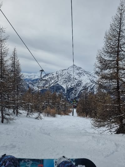 The image presents a first-person perspective from a snowboarder on a snow-covered ski slope in Cesana Torinese, Italy. In the immediate foreground, the front portion of a blue and black snowboard is visible, with two dark, patterned snowboard boots secured to its bindings. The ski slope directly ahead is covered in white snow, displaying tracks and minor undulations.
The midground features a ski run bordered on both sides by numerous coniferous trees with bare, dark branches. A ski lift system is operational, with cables extending through the upper frame. One empty ski lift chair is visible on the left side, suspended from a cable. Further down the slope on the right, another ski lift chair is descending, containing two individuals wearing bright green and yellow jackets. A black ski lift tower supports the cables in the lower mid-right.
The background is dominated by snow-capped mountain ranges. A prominent, jagged peak, largely covered in white snow with exposed dark rock, stands centrally. Additional distant mountains fade into the horizon under a uniformly overcast, light grey sky.