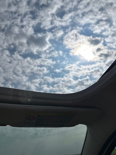 The image captures a view through a car's sunroof on a cloudy day. The sun is visible through breaks in the scattered clouds, casting a bright glow. The sunroof itself is visible in the foreground, with its interior trim and a warning label attached to the visor. The warning label contains text in Italian and several pictograms, including one depicting a person with a seatbelt and another showing an open book. The overall impression is one of travel, with the vast sky and clouds suggesting an open road. There are no people or specific landmarks visible that would allow for identification of the city of Longueil, France.