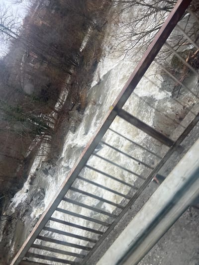The image depicts a raging river viewed from a bridge or overpass. The water is turbulent and white, suggesting a high flow rate, possibly due to recent rainfall or snowmelt. A metal railing with vertical bars is visible along the edge of the bridge, creating shadows on the water's surface. Trees with bare branches line the riverbank. A metal crash barrier is also visible. The location is Касапите, Bulgaria.