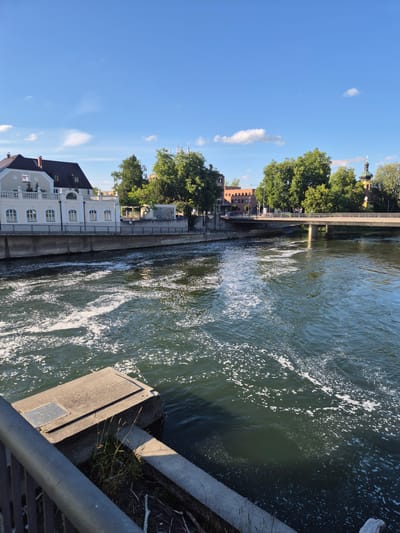 The image shows a view of the Isar river in Landshut, Germany, on a clear, sunny day. The river is flowing with white foam on its surface, suggesting some turbulence or rapids. On the left bank, there is a light-colored building with a dark roof, featuring arched windows and decorative white railings. Adjacent to it, a concrete wall separates the building from the river.
Across the river, a bridge with concrete pillars spans the water, carrying traffic. Beyond the bridge, trees and a building with an orange-red brick facade are visible. Further in the distance, a church with a green dome and a yellow tower stands out against the blue sky. The sky is clear with a few scattered white clouds. In the foreground, a dark metal railing is partially visible, along with a concrete structure jutting into the river.