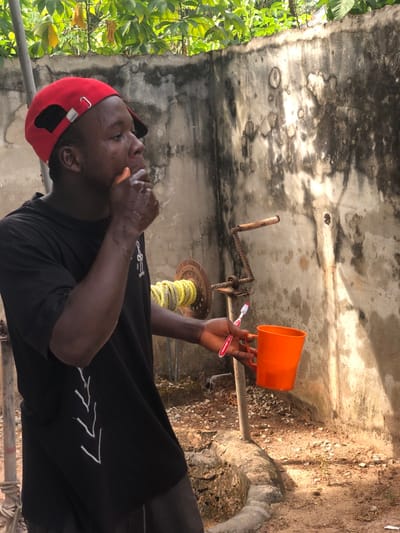 The image depicts an outdoor scene in Oraukwu, Nigeria. A male individual, wearing a red baseball cap and a black t-shirt, is positioned centrally. His right hand holds a small orange object to his mouth, with visible moisture on his chin and left cheek. His left hand simultaneously holds a white and red manual toothbrush and an orange plastic cup. In the background, a distressed concrete wall shows extensive dark staining and texture variations, with green foliage visible above its top edge. To the subject's right, a manually operated water well mechanism stands, featuring a rusted metal wheel wrapped with yellow rope and an accompanying crank handle. The ground beneath is an uneven surface composed of dirt and small gravel, illuminated by bright sunlight.