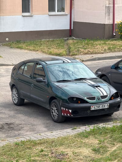 A dark green Renault Megane is parked on a paved surface, in front of a building with light peach and brown accents. The car features racing stripes on its hood and roof, and the front bumper has a shark-tooth design and modified headlights. A license plate reads "6295 BX-5" with "BY" indicating Belarus. To the right of the Megane, a portion of a darker vehicle is visible. The environment includes a sidewalk, a patch of dry grass, and a building with windows and downspouts. This scene is located in the city of Zhodzina, Belarus.