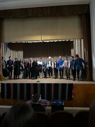 A stage performance is captured in a photograph taken in Voiniagovo, Bulgaria. The stage has a brown curtain backdrop and floor coverings with colorful stripes. A group of approximately fifteen people are standing in a line on the stage, dressed in what appears to be period or traditional costumes. The costumes include red fez hats, military-style jackets, dresses, and vests. An audience is partially visible to the right of the stage, with some viewers holding phones. A sound system or speaker is also visible in the background.
