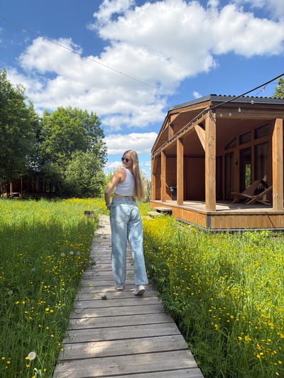 A young woman is captured posing on a wooden walkway in a bright, natural outdoor setting, likely in Vozhoiskii, Russia. She stands mostly sideways to the camera, with her head turned to look directly at it, displaying a slight smile. She has long, straight light-colored hair and is wearing a white sleeveless crop top, light blue baggy jeans, and sunglasses.

The wooden walkway stretches into the distance, bordered by a dense field of vibrant green grass profusely covered with small yellow flowers, possibly buttercups or similar wildflowers. Beyond this floral expanse, a line of large green trees with full canopies forms the background on the left side of the image.

To the woman's left, a rustic wooden building, resembling a cabin or gazebo, stands with a dark roof. It features a covered porch-like area with wooden posts and visible window panes. String lights with clear, round bulbs are strung along the eaves of the building, adding a decorative touch. A wooden chair is discernible inside the structure.

The scene is bathed in bright daylight under a clear blue sky dotted with scattered white, puffy clouds. The overall mood is pleasant and idyllic, suggesting a serene and relaxed environment. No visible text is present in the image.