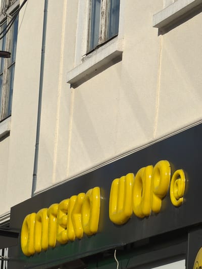 The image shows a close-up of a yellow, three-dimensional sign mounted on a dark background. The sign appears to be made of a glossy material and is shaped like a coiled object, possibly representing a stylized lightbulb or a spring. Several segments of the coil are visible, stacked vertically and curving outwards. The sign is partially obscured by the building's facade, which is a light-colored, textured material, possibly stucco. A portion of a window frame with weathered wood is visible on the left side of the image. The lighting suggests it is daytime, with bright sunlight casting shadows. The location context is Stryi, Ukraine. No people are visible in the image, and no specific activity or event is depicted. The overall impression is an exterior view of a building with signage.