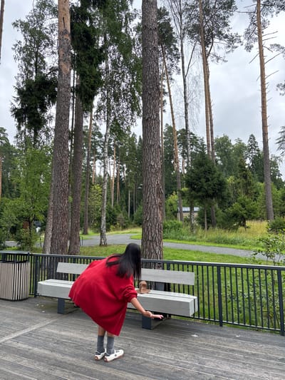 A young girl with long dark hair, wearing a vibrant red oversized sweater, grey socks, and white sneakers, is crouched on a wooden deck, reaching towards a light grey bench with a black metal railing. She appears to be interacting with something on or near the bench. To her left, a grey trash receptacle with vertical slats stands on the deck. The bench and railing overlook a grassy area with a gravel path winding through a dense forest of tall pine and birch trees. The sky is overcast. The scene is likely in a park or recreational area.