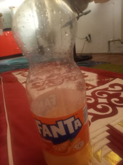 In an indoor setting, a partially filled plastic bottle of Fanta soda is prominently featured in the foreground, tilted to the right. The bottle is covered in condensation, indicating it is cold. The Fanta logo, with its distinctive orange and blue colors, is visible on the label. The liquid inside is a pale orange color. The bottle rests on a red and white patterned rug, which is laid over a wooden floor. In the background, a white wall is visible, with a black electrical outlet and a pipe. To the left, a glimpse of a red cabinet can be seen. The image appears to be taken from a slightly elevated angle, looking down towards the bottle. The overall environment suggests a casual indoor space, likely a home. The image does not provide any specific clues to its location within the city of Талапкер, Kazakhstan.