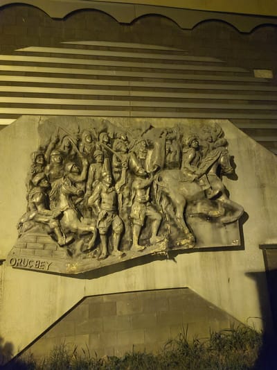 The image shows a low-relief bronze sculpture depicting a historical battle scene, likely a procession or charge, with many figures and a horse. The sculpture is mounted on a textured concrete wall, which is part of a larger architectural structure. Above the sculpture, there are horizontal bands of alternating light and dark materials, forming a decorative frieze. The inscription "ORUC BEY" is visible at the bottom of the sculpture. The scene is illuminated by artificial light, casting shadows and highlighting the metallic sheen of the bronze. In the foreground, there is a section of a grey, block-like structure and some sparse vegetation. The overall impression is of a public monument or memorial, likely in a city square or along a prominent building. The city of Osmangazi, Turkey, is known for its historical sites, and this sculpture may represent a significant event or figure from its past, possibly related to the Ottoman Empire.