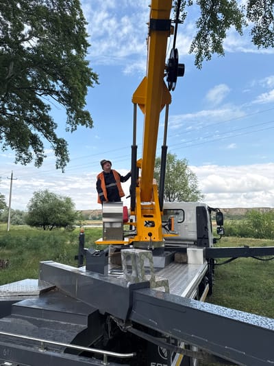In Kvemo Khvedureti, Georgia, a man wearing an orange reflective vest and a hat stands on a yellow crane attached to a white truck. The crane's arm is extended upwards, and the man appears to be operating or inspecting it. The truck is parked on a grassy area with trees and a blue sky with clouds in the background. The scene suggests an outdoor work setting, possibly for construction or maintenance.