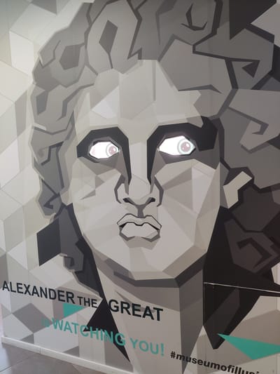 The image shows a large, abstract mural of Alexander the Great's face, rendered in a geometric, low-polygon style with shades of gray and white. The mural features two illuminated eyes that appear to be lit from behind, giving them a glowing effect. Below the face, text reads "ALEXANDER THE GREAT IS WATCHING YOU!" and "#museumofillusion". The mural is part of an exhibit at the Museum of Illusions in Thessaloniki, Greece, located at 13a Tsimiski Street. The mural is displayed on a wall, and the bottom edge of the image shows a white baseboard and a tiled floor.