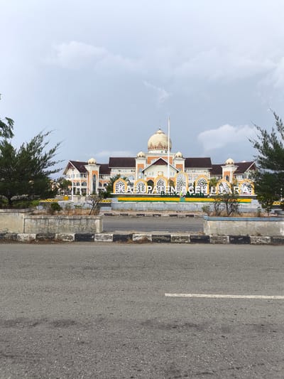 The image shows a grand, multi-story building with distinctive domed roofs and ornate architectural details, set against a cloudy sky. In front of the building, a large, decorative sign spells out "BUPATI ACEH UTARA" in prominent white lettering, surrounded by intricate yellow and green landscaping. The building appears to be a significant public structure, likely a government administrative building. The scene is viewed from across a paved road, with a concrete median strip separating the road from the building's grounds. The median is bordered by a low concrete wall with black and white painted stripes. Evergreen trees flank the building on both sides. The overall impression is of a well-maintained public space.