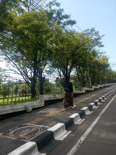 A person walks along a sidewalk next to a road. The sidewalk is made of grey paving stones with a strip of textured yellow stones. A black and white curb separates the sidewalk from the road. The road has a white line painted on it. A black metal fence runs along the other side of the sidewalk, separating it from a grassy area with trees. Overhead are trees and power lines. The location is not Khe Luang, Laos.