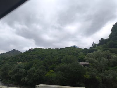 A wide, low-angle shot captures a verdant hillside under a cloudy sky. Dense, dark green trees cover the lower slopes, with lighter green foliage on the upper reaches of the hill. Several small buildings, appearing to be houses, are nestled on the top of the hill. The sky is overcast with heavy, gray clouds, obscuring any direct sunlight. In the foreground, a concrete barrier runs horizontally across the bottom of the frame. To the left, the edge of a dark, possibly car interior, frame is visible at the top. The scene suggests a rural or semi-rural setting in Armenia.