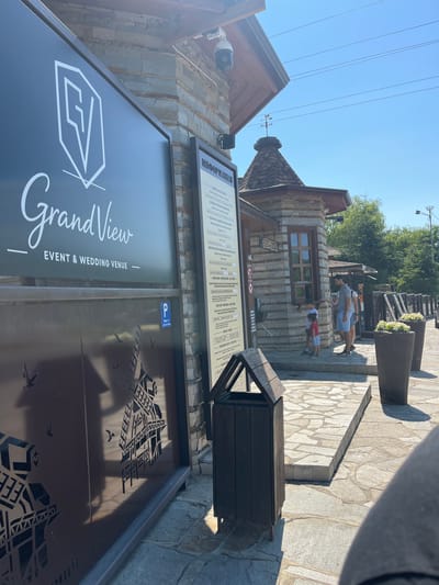 This image depicts the entrance area of GrandView Event & Wedding Venue in the city of Gorna Malina, Bulgaria. The scene is outdoors under a clear blue sky on a sunny day.
In the foreground, a large, dark sign with the "GrandView" logo and "EVENT & WEDDING VENUE" text is prominently displayed on the left. Below this sign, a decorative metal gate features cut-out silhouettes of windmills and birds. To the right of the main sign, a smaller information board with text in a foreign language is attached to a stone-built structure.
The stone-built structure appears to be a small kiosk or ticket booth with a wooden-shingled roof and a weather vane on top. A security camera is visible mounted on the overhang of the building. A dark wooden trash receptacle stands in front of the information board.
Several people are interacting near the kiosk. A man in a grey t-shirt and blue shorts is speaking to two young boys. One boy, wearing a red shirt and blue shorts, is standing closer to the kiosk window, while another boy in a white shirt and blue shorts is slightly behind him. Another individual, partially obscured, appears to be behind the man.
The ground is paved with irregular stone tiles. Large, dark planters filled with white flowers are situated to the right of the kiosk, and a wooden fence runs along the side of the pathway. The overall environment suggests a recreational or event-oriented location.