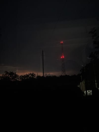 The image shows a telecommunications tower with red lights visible in the distance. The scene is dark, with the sky appearing to be twilight or night. Trees and power lines are also visible. The location is specified as Ганчарскі сельскі Савет, Belarus.