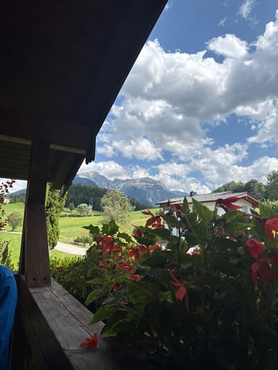This image captures a scenic view from a covered balcony in Bischofswiesen, Germany, on a cloudy but bright day. In the foreground, a wooden railing is visible with some red flowers and green foliage. To the left, a dark wooden beam supports the roof of the balcony, and a hint of a person in a blue shirt can be seen. Beyond the balcony, a rolling green landscape unfolds, dotted with trees and a winding road. In the middle ground, a white house with a dark roof stands nestled amongst the trees. The background is dominated by majestic mountains, their peaks partially obscured by fluffy white clouds against a clear blue sky. The overall scene evokes a sense of peaceful rural life amidst stunning natural beauty.