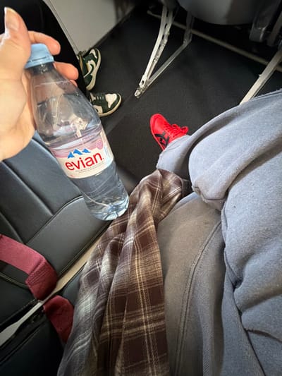 The image depicts an interior cabin, consistent with a vehicle or aircraft, situated in Villeneuve-le-Roi, France.

The scene focuses on a seated individual's lower body and a hand holding a bottle of water. The subject's light-skinned right hand grasps a clear 1-liter plastic bottle of Evian natural mineral water, which has a light blue cap and a distinctive white, red, and blue label; the bottle is approximately half-full. The individual wears light gray sweatpants and has a brown and white plaid blanket or shirt draped over their lap and legs. A red athletic shoe with black stripes, characteristic of an Onitsuka Tiger Mexico 66 style, is visible on their right foot.

On the dark-colored floor in front of the primary subject, a pair of green and white athletic shoes, consistent with a Nike Dunk Low style, indicates the presence of a second individual. The environment consists of dark grey or black padded seating with visible maroon seatbelt straps, a dark floor covering, and light grey metallic structures, likely seat frames or floor mounting rails. The overhead space and background beyond the immediate seating area are not clearly visible.