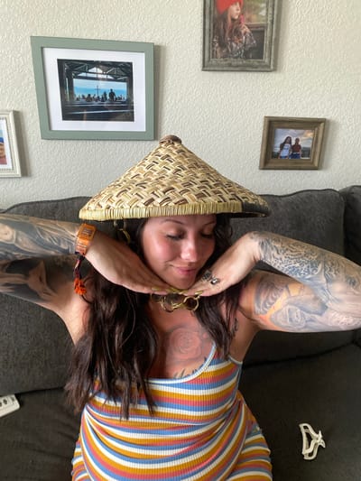 A woman with extensive tattoos on her arms and chest is seated on a dark sofa indoors. She is wearing a straw conical hat and a multicolored striped tank top. Her hands are placed on her chin, and she is looking down with her eyes closed. There are several framed photographs on the wall behind her, depicting people and landscapes. The scene is illuminated by ambient indoor lighting. The location is not specified in the image or its context.