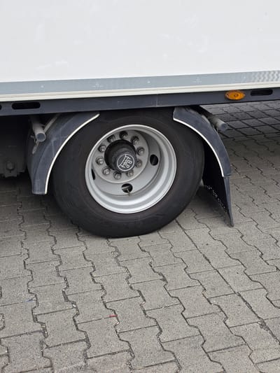 A close-up, eye-level, medium shot captures the rear wheel of a white truck parked on a cobblestone surface in Zeppelinheim, Germany. The wheel features a silver rim with multiple lug nuts and a black hubcap bearing the logo of a company with the letters "BP" inside a circular emblem. The tire is a Goodyear brand, size 235/75R17.5. The truck's body is visible in the upper portion of the frame, with a horizontal gray stripe along its lower edge. A black mudguard with a white trim partially covers the wheel, and a black plastic mudflap extends downwards from it. An orange reflector is visible on the side of the truck above the wheel. The ground is paved with interlocking gray cobblestones.