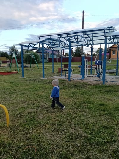 In Smolino, Russia, a young child wearing a grey hat, blue hooded sweatshirt, and dark pants walks across a grassy area. The child is moving from left to right in the frame. Behind the child, a covered outdoor gym and playground area is visible. This area features blue metal support structures and a white translucent roof. Within the gym area, various exercise machines are present, including what appears to be a leg press machine and a chest press machine. A sandbox with red trim and a swing set are also visible in the background, as well as several wooden houses with dark roofs. The ground is covered in green grass, and the sky is overcast with clouds.