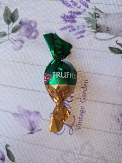 This media file is a top-down, eye-level, close-up shot of a single chocolate truffle in green and gold foil wrapping. The truffle is centrally placed and appears to be the sole focus of the image. The background is a textured, white fabric surface with a subtle floral pattern in shades of purple and green, featuring depictions of lavender, butterflies, and leaves. The text "TRUFFLE" is clearly visible in white lettering on the green portion of the foil, with smaller text "with nu" beneath it. To the left of "TRUFFLE," there is a red and gold circular logo with some smaller text. To the right of the truffle, the words "Vintage Garden" are printed vertically in purple script. The lighting suggests it is daytime, likely indoors, given the consistent illumination and lack of harsh shadows. There are no people or animals visible. The overall impression is one of a simple product presentation against a decorative backdrop, possibly for marketing or personal documentation. The location context of Kladno, Czechia, is noted but not visually evident in the image itself.