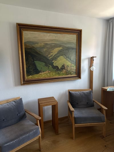 In St. Märgen, Germany, a living room scene features a large, framed oil painting of a mountainous landscape hanging on a textured white wall. Below the painting, two wooden armchairs with dark gray cushions are positioned on a polished wooden floor. Between the chairs sits a small, slatted wooden side table. To the right of the right armchair, a tall, narrow wooden floor lamp with a single exposed light bulb stands near a light gray curtain. A wooden desk is visible to the far right, with a magazine or booklet resting on it. The overall atmosphere is that of a quiet, indoor space.