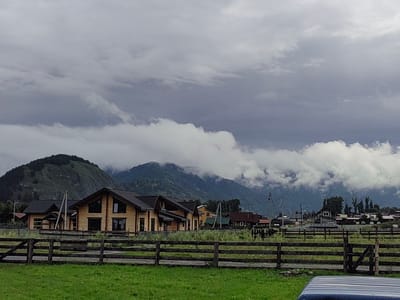 The image depicts a rural landscape in Anos, Russia, under a cloudy sky. The foreground features a green grassy field with a weathered wooden fence. Behind the fence, a cluster of modern wooden houses with dark roofs are visible, suggesting a residential area. In the background, a mountain range is shrouded in mist and low-hanging clouds, creating a dramatic and somewhat somber atmosphere. The overall scene conveys a sense of natural beauty and quietude.