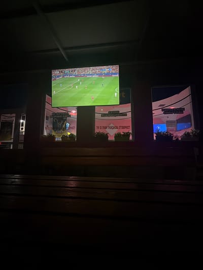 The image depicts an outdoor scene at night, likely in a public establishment in Ognianovo, Bulgaria. Dominating the view is a large television screen displaying a live football match. The screen shows the score as 0-0, with the teams POR and ESP indicated. The game is in its 32nd minute. Advertising for "Qatar Airways" is visible on the screen.
Below the large screen, two illuminated rectangular displays are visible through windows. The left display features Cyrillic text that translates to "SUPER COEFFICIENTS" and "CITIZEN RESPONSIBILITY." A graphic of a muscular figure is also present. The right display shows Cyrillic text "VIRTUAL SPORTS" and appears to be an advertisement for a betting or gaming establishment.
The foreground is dominated by a dark wooden bench, suggesting a seating area for patrons. The overall lighting is dim, with the main light sources being the screens and some interior lighting visible through the windows. The environment appears to be a commercial area, possibly a cafe or bar, where people would gather to watch sports. The presence of potted plants beneath the illuminated displays adds a touch of decoration.