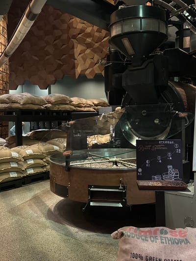 This image depicts the interior of a coffee roasting facility, likely located in the Jing'an District of China. In the foreground, a large coffee roasting machine with a copper-colored drum is visible, filled with coffee beans. A glass display case protects the beans. To the right, a chalk-style sign titled "The STARBUCKS Roast Spectrum" illustrates different stages of coffee roasting, from "Yellow Bean" to "Dark Roast," with accompanying Chinese text. Behind the roaster, stacks of burlap sacks filled with coffee beans are neatly arranged on shelves and the floor. One sack in the lower right corner is labeled "PRODUCE OF ETHIOPIA 100% GREEN COFFEE." In the background, a wall is decorated with a geometric, honeycomb-like pattern made of wood. Industrial pipes and other machinery are also visible, suggesting a working production environment. The lighting is somewhat dim, creating shadows and highlighting the textures of the coffee beans and sacks.