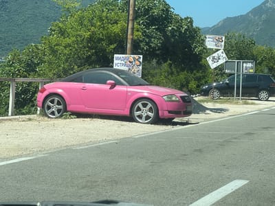 A bright pink convertible Audi TT is parked on a gravelly shoulder next to a paved road. The car has a black soft-top roof and silver five-spoke rims. Behind the car, lush green trees and a steep, vegetated mountainside form the backdrop. The scene is bathed in bright sunlight, suggesting a clear day, likely in the late morning or early afternoon.
A utility pole stands near the car, with several signs attached. One sign, featuring images of mussels and oysters, has Cyrillic text that reads "МИДИИ УСТРИЦЫ". Another sign below it is tilted and displays a black and white graphic. Further to the right, a black Volkswagen Golf is parked. Beyond the Golf, a larger sign is visible, appearing to be a towing service advertisement, with the words "TOWING SERVICE" and a phone number.
The overall impression is a roadside scene in a scenic, possibly coastal, mountainous region, likely in Montenegro, given the location context. The vibrant pink of the car contrasts with the natural greenery and the other vehicles present. No people are visible in the frame.