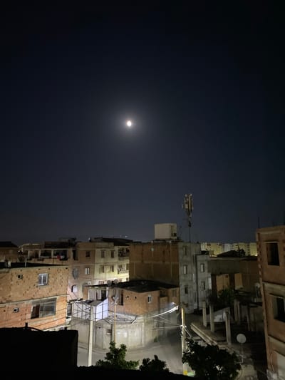 This nighttime photograph depicts a densely built residential area in Baraki, Algeria. The upper half of the image is dominated by a dark, clear night sky, with a bright, full moon prominently visible in the upper left quadrant, casting a soft glow. A few faint stars can be discerned scattered across the sky.
The lower and right portions of the frame are filled with multi-story buildings, characteristic of an urban environment. These structures showcase a mix of construction materials, including exposed red brick and weathered concrete, with numerous windows, balconies, and air conditioning units visible. Satellite dishes are attached to several buildings. In the mid-ground, a tall mast or antenna structure with multiple components rises above the rooftops. Utility poles and overhead electrical wires crisscross between the buildings. Portions of a street or alleyway are visible at the ground level, illuminated by artificial lights. A dark, silhouetted tree stands at the far right edge of the image.
No people or specific activities are visible, conveying a sense of stillness and quiet typical of a residential area at night. There is no visible text in the image.