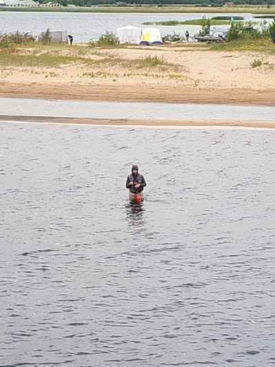 A lone individual stands in the choppy water of a body of water, facing towards the right side of the frame. The person is wearing a dark hooded jacket and waders that extend up to their mid-thigh, with a bright orange object visible at the waist area. The background shows a sandy shore with sparse vegetation. Further back, several tents and a small boat are visible on the shore. The overall environment suggests an outdoor recreational area, potentially near a campsite or a fishing spot in Shalsky, Russia.