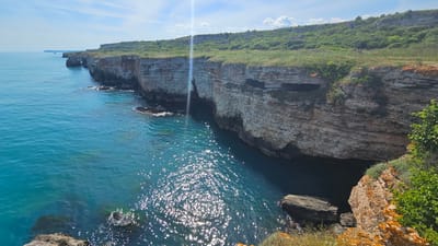 A picturesque view of the Kamen Bryag coastline in Bulgaria unfolds, featuring a series of layered limestone cliffs topped with green vegetation. The teal-colored sea extends to the left, dotted with sun glares. The cliff faces reveal horizontal strata. Erosion has created caves and rocky formations at the base.