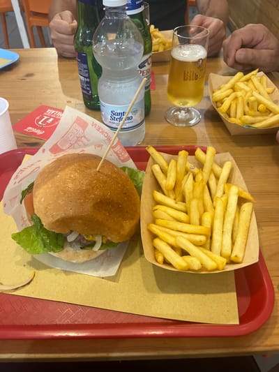 In this medium shot, a wooden table is laden with food and drinks. Two people's hands are visible on the left side of the frame. In the foreground, a hamburger sits on a red tray lined with brown paper. The hamburger has a fluffy bun, green lettuce, and other toppings. Beside it, a bottle of "Santo Stefano" mineral water and a green bottle are placed. A glass of beer, with a prominent "Bitburger" logo, is also on the table, next to a second empty glass. A portion of french fries in a paper container is positioned in the upper portion of the frame, while another portion of fries sits on a separate brown paper wrapper. The scene appears to be a casual meal, possibly at a restaurant or fast-food establishment, given the paper packaging and branding elements like "Coca-Cola" printed on the burger wrapper. The lighting suggests it could be daytime. The overall atmosphere is relaxed and informal. Visible text includes "PILLATA", "HATA PER ESMERE", "1", "Coca-Cola", "9% Vol", "ETO SU", "ESCLUS", "EFONINASE", "QUALITARI", "PER", "MAST", "20", "Santo", "ACQUA OLI MINERALE", "NAT", and "Bitburger". The location context provided indicates Vallo della Lucania, Italy.