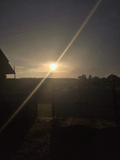This image depicts a landscape scene, likely in Малая Уса, Russia, during what appears to be sunset or sunrise, given the bright, hazy sun low in the sky. The sun is the primary subject, casting a strong lens flare that diagonally bisects the image.
The scene is divided into two main areas by the horizon. On the left, a hazy sky dominates, with the bright sun positioned towards the center. Several power lines are visible as thin, dark lines against the sky, suggesting an outdoor, possibly rural or semi-rural, setting.
The right side of the image is darker, featuring silhouetted elements. A treeline forms the distant boundary of the landscape. Closer to the foreground, there's a dark, indistinct structure, possibly a building or roofline, in the lower left. On the right side of the frame, a darker, textured area suggests a fence, hedge, or wall, with some lighter patches that could be illuminated by the faint light. A square, lighter section within this darker area might be a wooden structure, like a shed or a section of fencing.
The overall mood is somewhat atmospheric and potentially serene, due to the natural light of dawn or dusk. The presence of power lines hints at human infrastructure within the natural environment. No people or text are visible in the image. The time of day is clearly indicated by the low sun and the quality of light, which is soft and diffused, suggesting either early morning or late evening.