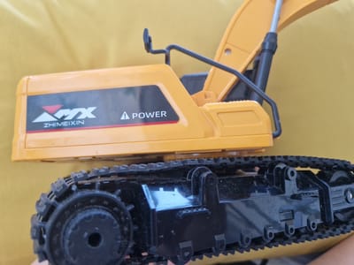 A close-up, overhead shot shows the upper body and tracked chassis of a yellow toy excavator. The excavator has black tracks and a bright yellow body with "AMX ZHIMEIXIN" and a triangular logo on its side, as well as a "POWER" label. The excavator is positioned on a textured yellow surface. There are no people present in the image. The setting is not identifiable as being in Fresnes, France.