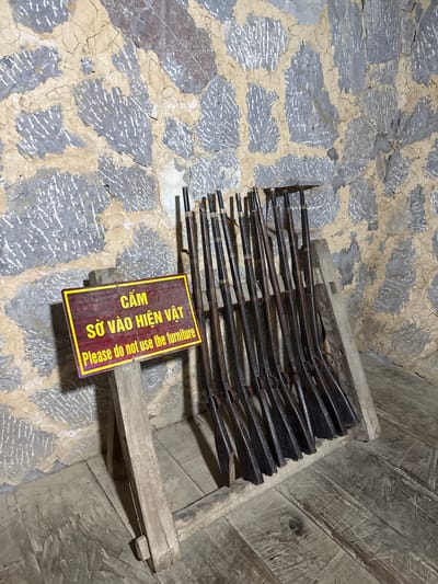 The image shows a collection of antique rifles displayed on a rough wooden rack in what appears to be a museum or historical site. The walls are constructed of stone, and the floor is uneven and textured. A sign in Vietnamese and English reads "Please do not use the furniture," suggesting that the rifles and rack are part of an exhibit. Based on the metadata provided, this image was taken in Sà Phìn Commune, Vietnam.