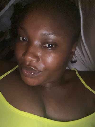 A close-up, frontal portrait photograph captures a dark-skinned adult female subject in an indoor setting within Dzhidu, Nigeria. The subject is wearing a neon yellow camisole, visible across her shoulders and upper torso. Her lips are slightly parted and appear glossy. Her gaze is directed towards the camera. The background is indistinct and dimly lit, suggestive of low-light conditions. No other individuals or specific objects are discernible.