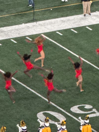 The image displays an artificial green turf sports field in East Rutherford, United States, marked with white lines including a broad solid stripe, dashed segments, and a large curved letter 'C' or 'G'. Six female individuals are prominent in the central foreground, all barefoot and clad in identical red one-piece leotards. They are engaged in dynamic, synchronized dance movements, with poses indicating high leg extensions, arm raises, and various mid-motion actions. Five of these performers exhibit dark complexions and dark hair, while one has a lighter complexion and light brown hair. In the upper background, two male figures are partially visible: one wearing tan trousers and a dark shirt, and another in blue jeans and a dark shirt positioned near a black microphone stand. In the lower foreground, multiple individuals in marching band uniforms are present, featuring yellow/orange tunics, dark trousers, and tall hats adorned with yellow plumes. Some of these individuals appear to be holding brass instruments.