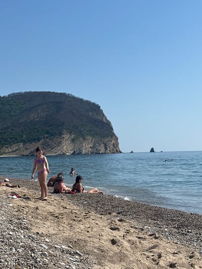 The image depicts a sunny day at a beach, likely in Đurovići, Montenegro. The scene is divided horizontally: the top portion shows a clear, bright blue sky, transitioning to the shimmering blue water of the Adriatic Sea. The bottom portion of the image is dominated by a sandy and pebbly beach, with a large, tree-covered mountain rising dramatically on the left side, bordering the sea.
Several people are visible enjoying the beach. In the foreground, a woman in a patterned bikini stands on the sand, facing away from the camera. Further back, a group of people are relaxing and interacting on the shore. Some are seated, while others appear to be in the water. One person is in the process of diving or jumping into the water, with their body partially submerged. The presence of people in swimwear and the overall relaxed atmosphere suggest a warm, pleasant day suitable for swimming and sunbathing.
The water appears calm with gentle waves lapping at the shore, and the sunlight creates a bright, summery ambiance. The texture of the beach, a mix of fine sand and small pebbles, is clearly visible. The mountain's dense vegetation suggests a lush, natural landscape. No visible text is present in the image. The overall impression is one of a leisurely day at a beautiful coastal location.