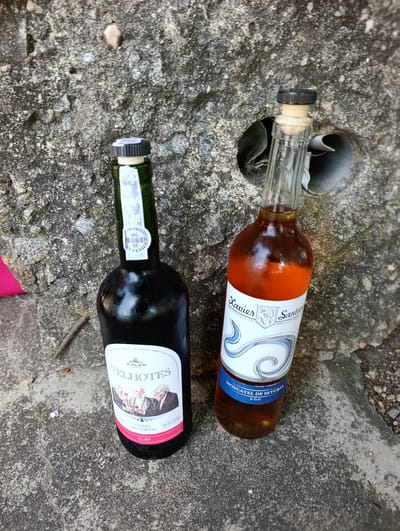Two bottles of wine are on a rough concrete surface. The bottle on the left is dark green and has a label with a blue and white design featuring three men. The label also reads "CALEM VELHOTE'S VINHO DO PORTO RUBY". A small sticker on the neck of the bottle has the text "XC 10648 05". The bottle on the right is clear glass and contains a reddish-brown liquid. Its label is white and blue and says "Xavier Santan", "EST 1920", and "MOSCATEL DE SETUBAL D.O.C.". The bottles are positioned near a circular opening in the concrete, which appears to be a drain or ventilation hole. The concrete surface is textured and appears to be outdoors. The image is taken from a high angle, looking down at the bottles. The location is identified as Trofa, Portugal.