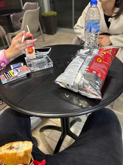 A high-angle shot reveals a black, round table laden with objects, surrounded by two individuals. One person, wearing black jeans, holds a half-eaten sandwich. On the table is a water bottle, a bag of chips, several packs of cigarettes, an ashtray, a phone, and a hand holding a lit cigarette. The scene takes place outdoors, with glimpses of tiled flooring and seating in the background. The cigarette packs feature brands such as Dunhill and Captain Black. No indication of Pirdop, Bulgaria.