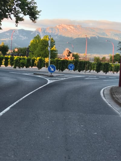 The image shows a road in Haro, Spain, with a mountain range in the background. The road is paved and has white lines indicating lanes. There are two blue traffic signs visible: one with a white arrow pointing down and to the left, and another with a white arrow indicating a roundabout. A row of neatly trimmed green hedges runs along the side of the road. In the distance, a rocky mountain range is illuminated by the warm light of what appears to be sunrise or sunset. The sky above is a soft blue with some wispy clouds. The overall scene suggests a peaceful setting on the outskirts of the city.
