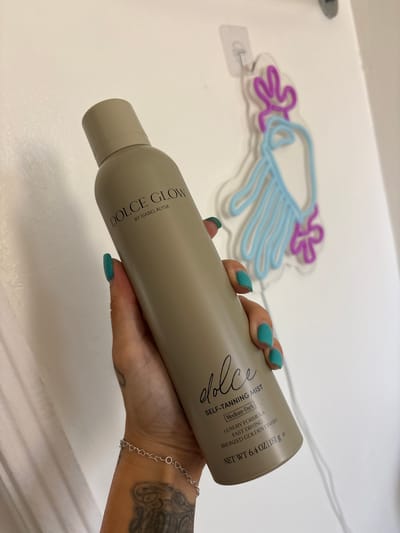 A close-up, indoor shot from Seal Beach, USA, features a person's hand holding a cylindrical bottle of self-tanning mist against a plain white background. The hand, adorned with teal nail polish, a delicate silver heart-link bracelet, and a visible tattoo on the forearm, grips the bottle.
The bottle, in a matte, light beige or taupe color, is angled across the frame. Prominent text on the bottle reads "DOLCE GLOW BY ISABEL ALYSA" vertically on one side, and "dolce" in a stylized script, followed by "SELF-TANNING MIST". Further details on the bottle specify "Medium-Dark", "LUXURY FORMULA", "FAST DRYING", and "BRONZED GOLDEN FINISH". The net weight is stated as "NET WT 6.4 OZ / 181 g e".
In the soft-focus background, a decorative neon sign shaped like a cloud with raindrops is visible. The cloud outline is purple, and the raindrops are light blue, mounted on a clear acrylic base. The overall setting appears to be a clean, bright indoor space, possibly for a product showcase or personal use.